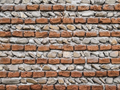 A wall constructed with alternating layers of red bricks and irregularly shaped gray stones, set in light gray mortar, creating a rustic and textured appearance.