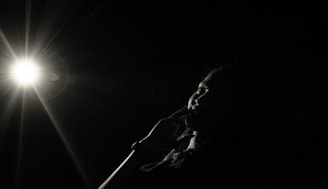 A person is partially visible in a darkened space, with a bright light source creating a dramatic flare effect. The light illuminates part of the face, highlighting a thoughtful or contemplative expression.