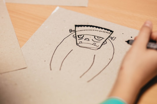 A hand is drawing a cartoon-like sketch of a simplified Frankenstein-style character on a plain sheet of paper. The character has a stitched square-shaped head with protruding ears and a blank expression. The surrounding area includes other sheets of paper on a wooden surface.