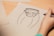 A hand is drawing a cartoon-like sketch of a simplified Frankenstein-style character on a plain sheet of paper. The character has a stitched square-shaped head with protruding ears and a blank expression. The surrounding area includes other sheets of paper on a wooden surface.