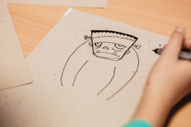 A hand is drawing a cartoon-like sketch of a simplified Frankenstein-style character on a plain sheet of paper. The character has a stitched square-shaped head with protruding ears and a blank expression. The surrounding area includes other sheets of paper on a wooden surface.