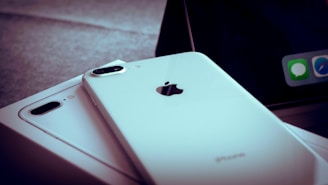 A close-up of two white iPhones, one of which is laying flat on a box. The Apple logo and camera lens are clearly visible. A portion of a screen with app icons is partially visible in the background.