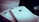 A close-up of two white iPhones, one of which is laying flat on a box. The Apple logo and camera lens are clearly visible. A portion of a screen with app icons is partially visible in the background.