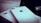 A close-up of two white iPhones, one of which is laying flat on a box. The Apple logo and camera lens are clearly visible. A portion of a screen with app icons is partially visible in the background.