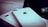 A close-up of two white iPhones, one of which is laying flat on a box. The Apple logo and camera lens are clearly visible. A portion of a screen with app icons is partially visible in the background.