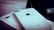 A close-up of two white iPhones, one of which is laying flat on a box. The Apple logo and camera lens are clearly visible. A portion of a screen with app icons is partially visible in the background.