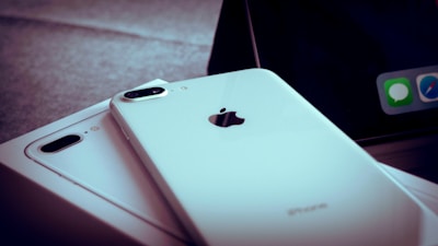 A close-up of two white iPhones, one of which is laying flat on a box. The Apple logo and camera lens are clearly visible. A portion of a screen with app icons is partially visible in the background.