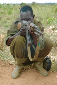 A person crouching in a field, aiming a rifle towards the camera. The individual is dressed in military attire and green rubber boots, focusing intensely. A water canteen is placed on the ground beside them, surrounded by grass and dry vegetation.