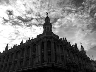 A dramatic black and white photograph of an ornate, historic building with an elaborate facade, featuring sculptures and intricate architectural details. The sky is filled with textured, partly cloudy formations.