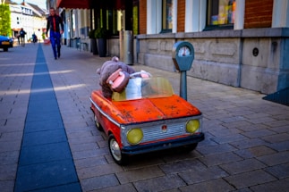 A small, vintage-style kiddie ride car with a stuffed hippopotamus toy inside is positioned on a cobblestone street next to a parking meter. In the background, a person walks along the sidewalk lined with storefronts.