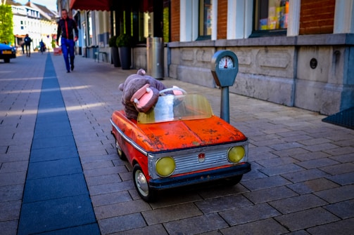 A small, vintage-style kiddie ride car with a stuffed hippopotamus toy inside is positioned on a cobblestone street next to a parking meter. In the background, a person walks along the sidewalk lined with storefronts.