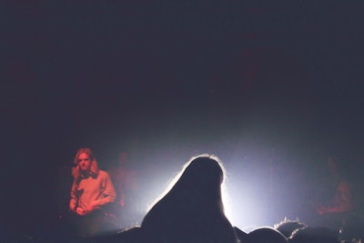 A stage performance with a focus on a person standing at the center with their back to the camera, illuminated by bright stage lights creating a silhouette. Another person is visible on the left side of the stage, partially lit in a subdued manner, suggesting a concert or music event setting.