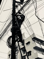 A person is climbing a ladder attached to a utility pole, surrounded by a complex web of cables and wires. The scene is set against the backdrop of buildings, creating a busy urban environment.