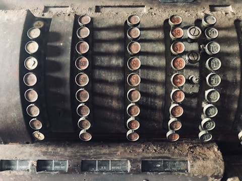 An old-fashioned, dusty cash register with multiple round keys displaying various monetary values. The keys are arranged in rows, featuring both numbers and words such as 'cash' and 'paid out.' The register has a worn, vintage appearance, indicating significant age and use.