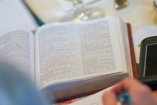 An open book with text printed in both black and red lettering, suggesting it's possibly a religious text or a study guide. There's a notebook nearby and a person's hand holding a pen, indicating someone is taking notes or studying.