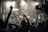 A lively concert scene with a large crowd facing a stage where several performers and a DJ are present. The audience has their hands raised in excitement, capturing the energetic atmosphere. The lighting is dramatic with spotlights emphasizing the performers.