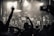 A lively concert scene with a large crowd facing a stage where several performers and a DJ are present. The audience has their hands raised in excitement, capturing the energetic atmosphere. The lighting is dramatic with spotlights emphasizing the performers.
