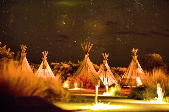 A row of illuminated teepees under a starry night sky with some visible greenery and vegetation in the foreground. The scene is warmly lit, suggesting a serene and tranquil outdoor setting.