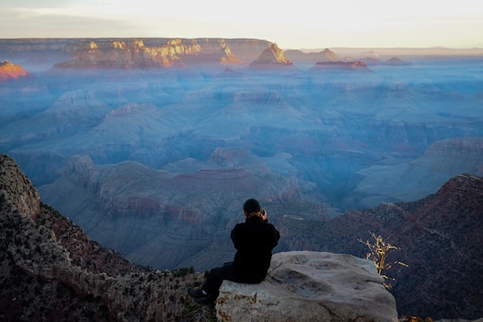 A person sitting on a large rock at the edge of a cliff, capturing a photo of a vast and colorful canyon landscape bathed in soft, early morning or evening light. The scene includes layered rock formations with shades of orange, pink, and blue, partially shrouded in mist, under a clear sky.