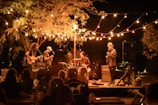 A nighttime outdoor concert featuring a band performing on a small stage. Strings of colorful lights drape overhead, creating a warm glow. The band consists of a drummer, a guitarist, a bassist, and a lead singer. An audience is present, enjoying the music under a large tree.