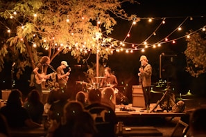A nighttime outdoor concert featuring a band performing on a small stage. Strings of colorful lights drape overhead, creating a warm glow. The band consists of a drummer, a guitarist, a bassist, and a lead singer. An audience is present, enjoying the music under a large tree.