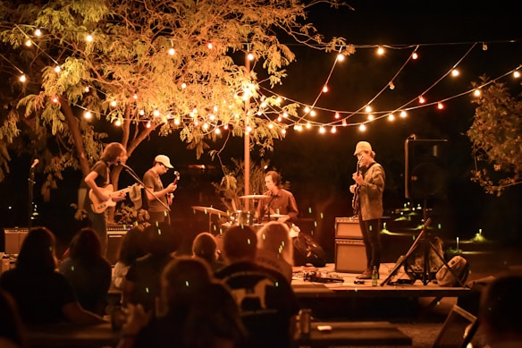 A nighttime outdoor concert featuring a band performing on a small stage. Strings of colorful lights drape overhead, creating a warm glow. The band consists of a drummer, a guitarist, a bassist, and a lead singer. An audience is present, enjoying the music under a large tree.
