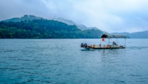 A traditional wooden boat with a canopy sails across a large, calm body of water. Misty hills covered in lush green trees form the background, creating a serene scene.