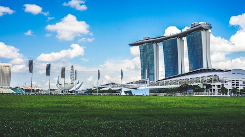 A modern urban landscape features a prominent, futuristic building with three towers connected by a skybridge. The foreground includes a vast, lush green lawn, and the sky is bright with scattered clouds.