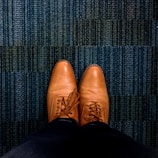 A pair of brown leather dress shoes is seen from an overhead perspective, resting on a textured blue carpet. The shoes are neatly laced and polished, showing detailed stitching and perforations.