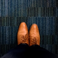 A pair of brown leather dress shoes is seen from an overhead perspective, resting on a textured blue carpet. The shoes are neatly laced and polished, showing detailed stitching and perforations.