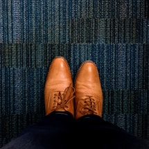 A pair of brown leather dress shoes is seen from an overhead perspective, resting on a textured blue carpet. The shoes are neatly laced and polished, showing detailed stitching and perforations.