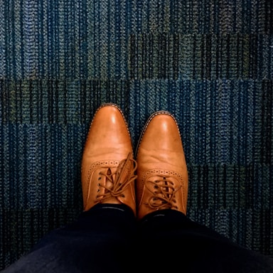 A pair of brown leather dress shoes is seen from an overhead perspective, resting on a textured blue carpet. The shoes are neatly laced and polished, showing detailed stitching and perforations.