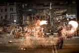 A street performer is entertaining a crowd with a fire show in what appears to be a public square. The performer is manipulating fire, creating a spectacle with sparks flying in the air. The audience is standing and sitting around, watching the act. There is a fountain in the background, and several buildings with lights are visible. There are also bicycles and other street elements.