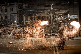 A street performer is entertaining a crowd with a fire show in what appears to be a public square. The performer is manipulating fire, creating a spectacle with sparks flying in the air. The audience is standing and sitting around, watching the act. There is a fountain in the background, and several buildings with lights are visible. There are also bicycles and other street elements.