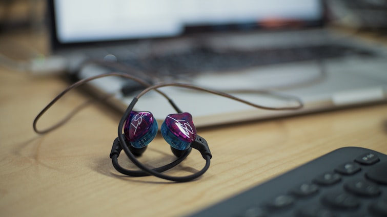 A pair of in-ear headphones with a black cord and purple-blue earpieces is lying on a wooden desk. A blurred laptop and keyboard are in the background, suggesting a workspace or office setting.