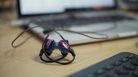 A pair of in-ear headphones with a black cord and purple-blue earpieces is lying on a wooden desk. A blurred laptop and keyboard are in the background, suggesting a workspace or office setting.
