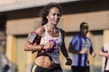 A woman is participating in a marathon, wearing a sports bra and shorts, with earphones connected to a device on her arm. Her focused expression and athletic posture suggest determination. Other runners are visible in the background, slightly out of focus.