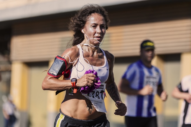A woman is participating in a marathon, wearing a sports bra and shorts, with earphones connected to a device on her arm. Her focused expression and athletic posture suggest determination. Other runners are visible in the background, slightly out of focus.