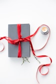 A neatly wrapped gray book adorned with a vivid red ribbon lies beside a lit tealight candle in a small holder. Sprigs of green foliage peek out from beneath the ribbon, adding a touch of nature to the composition. The white background enhances the contrast of the colors.