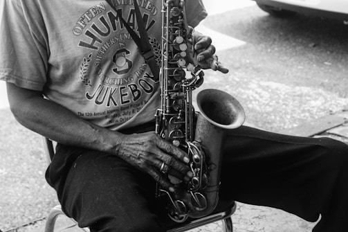 A close-up in black and white of a person sitting and playing a saxophone, focusing on the hands and instrument. The person is wearing a T-shirt with the words 'Human Jukebox' visible among other text. The texture of the saxophone and the lighting create a nostalgic feel.