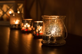 A series of glass candle holders are lined up on a dark wooden surface, softly illuminated by the warm glow of flickering tealight candles inside them. The glass is slightly frosted, creating a speckled light pattern that adds a cozy ambiance to the setting. The background is softly blurred, enhancing the focus on the candles and their gentle light.