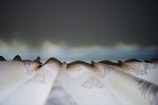 A close-up view of pleated curtains, possibly with intricate patterns, against a softly blurred background suggesting a twilight sky. The perspective focuses on the folds and details of the fabric.