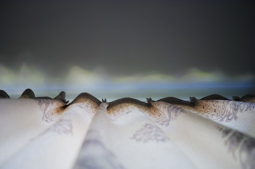 A close-up view of pleated curtains, possibly with intricate patterns, against a softly blurred background suggesting a twilight sky. The perspective focuses on the folds and details of the fabric.