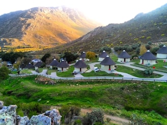A picturesque landscape featuring a series of round huts with thatched roofs arranged neatly in a valley. The huts are surrounded by green grass and are positioned along a winding path. In the background, a mountain range is illuminated by sunlight, creating a calm and serene atmosphere.