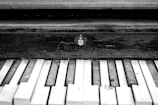An aged piano keyboard with worn and chipped keys against a wooden background. The name 'Wurlitzer' is inscribed, indicating the brand of the piano. The monochrome color scheme adds a classic and vintage feel.