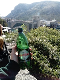 A plastic bottle labeled 'kiseljak' is held up by a hand, with a scenic background featuring a stone arch bridge and surrounding buildings and vegetation. Rolling hills and mountains rise in the distance under a clear sky.