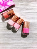 Four nail polish bottles with rose-gold caps are arranged on a light wooden surface. The shades of the nail polish include various tones of pink. A pink fabric with white designs is partially visible in the top left corner.