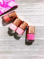 Four nail polish bottles with rose-gold caps are arranged on a light wooden surface. The shades of the nail polish include various tones of pink. A pink fabric with white designs is partially visible in the top left corner.
