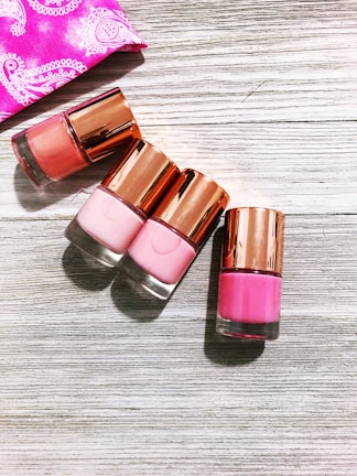 Four nail polish bottles with rose-gold caps are arranged on a light wooden surface. The shades of the nail polish include various tones of pink. A pink fabric with white designs is partially visible in the top left corner.