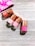 Four nail polish bottles with rose-gold caps are arranged on a light wooden surface. The shades of the nail polish include various tones of pink. A pink fabric with white designs is partially visible in the top left corner.